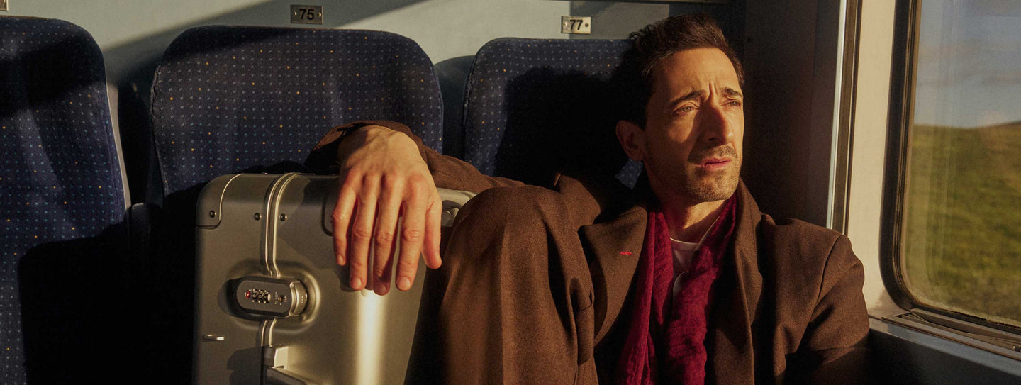 Adrien Brody sits by a train window, bathed in warm sunlight. He wears a brown coat and a deep red scarf, gazing outside with a contemplative expression. His hand rests on a Monos Aluminum Carry-On Plus suitcase in Aspen Silver beside him. The train’s blue seats and numbered labels 