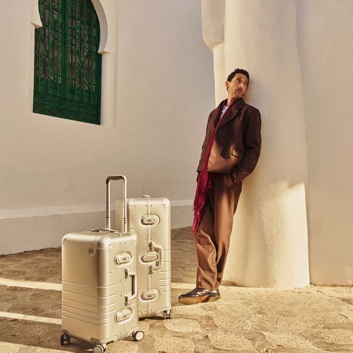 Adrien Brody leans against a curved white pillar, bathed in warm sunlight. Two Monos aluminum suitcases in Aspen Silver stand beside him on a textured stone path, with a green window and white architecture in the background.