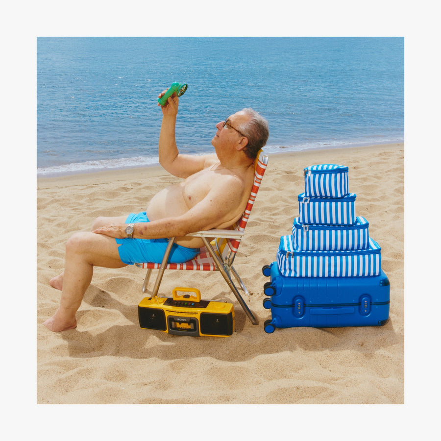 Jungle Green | Man relaxing on a chair, cooling off with a portable fan beside a stack of Coastal Blue Hybrid suitcases and Poplin Stripe Coastal Blue Packing Cubes in various sizes