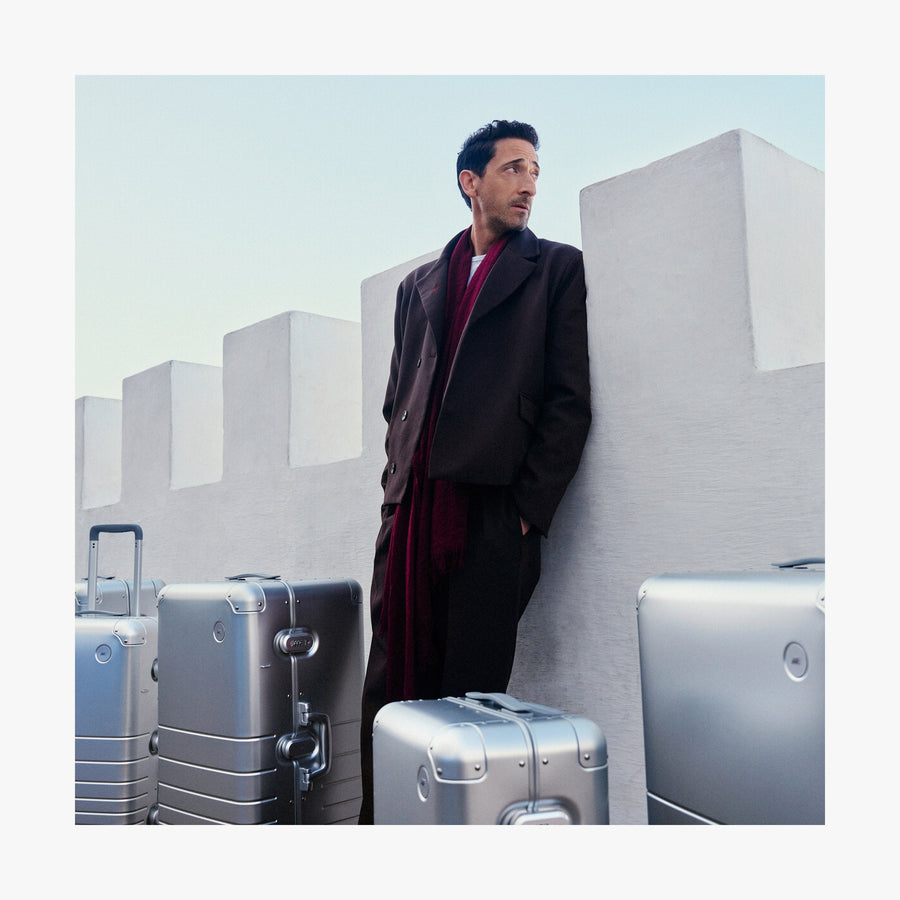 Champagne Gold | Adrien Brody leans against a white fortress-like wall, gazing into the distance, with multiple Monos aluminum suitcases in aspen silver around him.