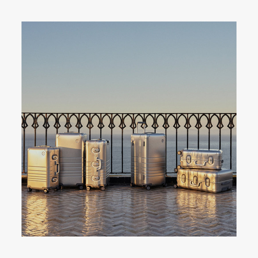 Aspen Silver | A collection of Monos aluminum suitcases in Aspen Silver in various sizes is arranged on a sunlit terrace with a wrought iron railing, overlooking the ocean in the background.