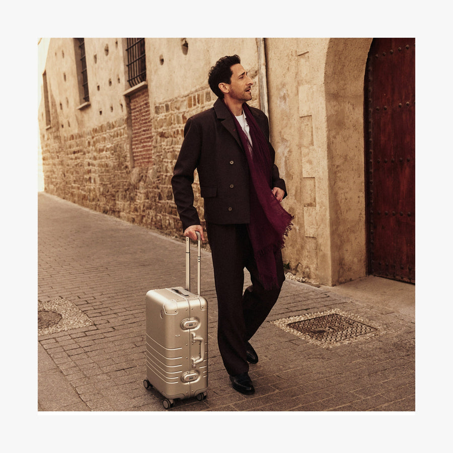 Champagne Gold | Adrien Brody dressed in a dark brown suit, white shirt, and a deep burgundy scarf walks down a historic cobblestone street while pulling a Monos Aluminum carry-on plus suitcase in aspen silver. The background features an old stone building with a wooden door, adding to the image's timeless, travel-inspired aesthetic.