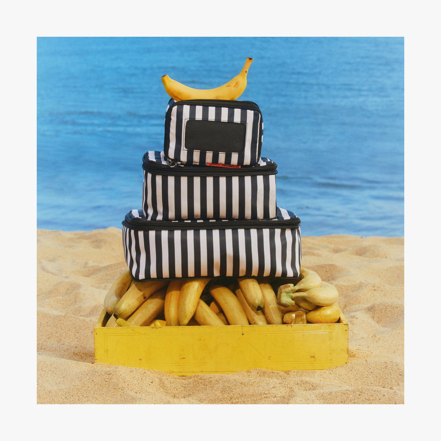 Set of Four / Poplin Stripe Black | Stack of Poplin Stripe Black compressible packing cubes arranged on a crate of bananas on a sandy beach, with a single banana placed on top