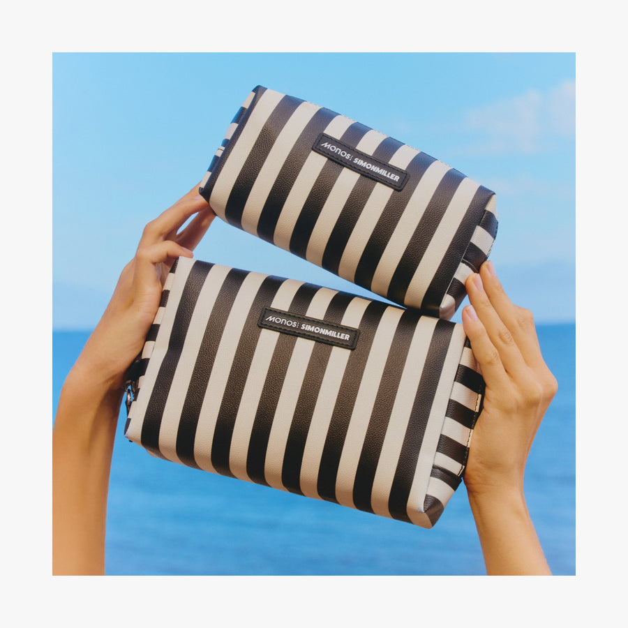 Large / Poplin Stripe Black | Two hands holding up the two sizes od Metro Toiletry Cases in Poplin Stripe Black against a bright ocean background