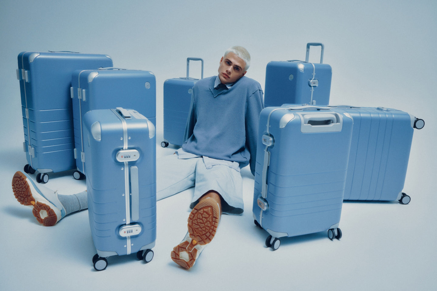 A model in a blue sweater sits among various sizes of Hybrid luggage in Elemental Blue on a light blue background.