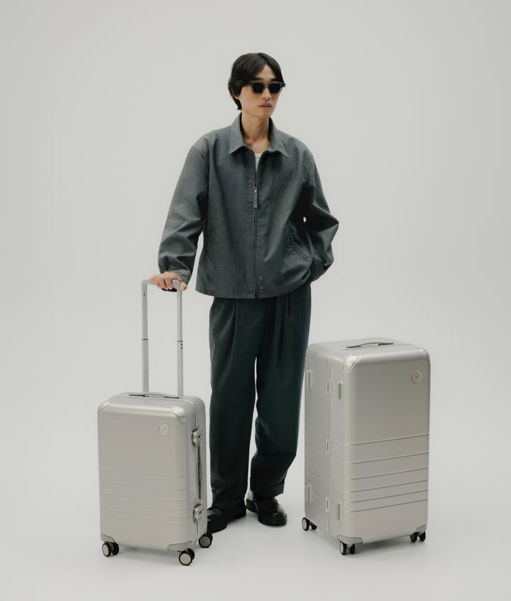 A man wearing a grey outfit and sunglasses stands beside the Monos Hybrid Carry-On and Hybrid Trunk in Silver. The minimalist travel set highlights Monos’ sleek aluminum design and modern craftsmanship.