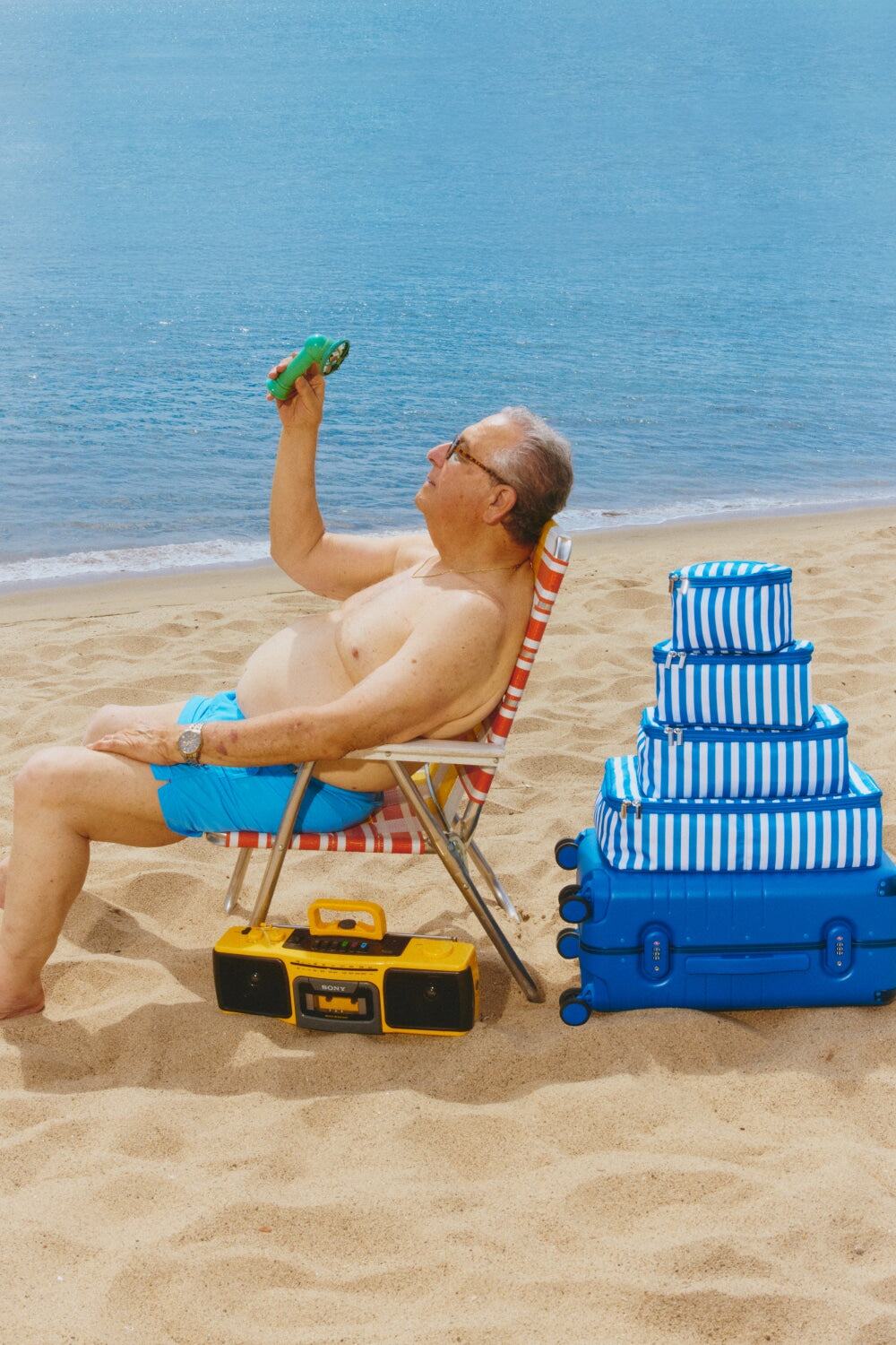 Man sitting on a beach chair with a blue suitcase and stack of packing cubes as well as a boombox, holding up a portable fan.