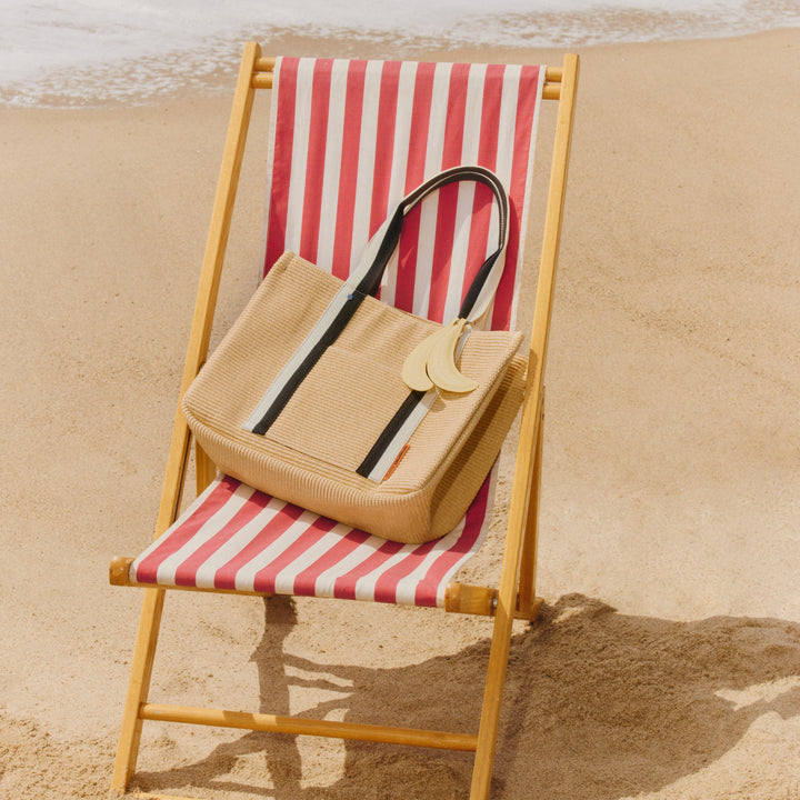 Straw handbag with black and white striped handle on a red and white striped beach chair by the ocean.