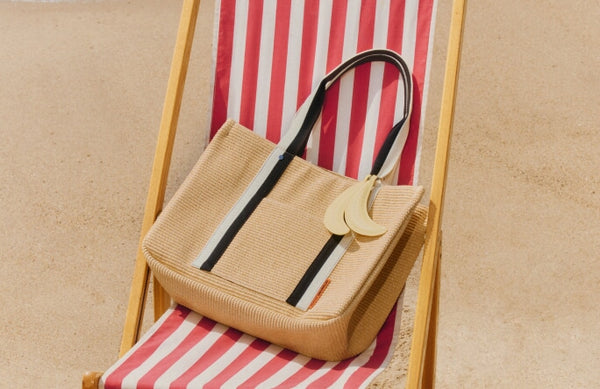 Straw handbag with black and white striped handle on a red and white striped beach chair by the ocean.