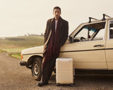 Adrien Brody stands beside a vintage cream-coloured car on a rural road, with a Monos aluminum carry-on plus in aspen silver by his feet and rolling green hills in the background.