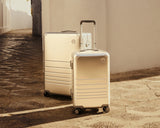 Two Monos aluminum suitcases in Aspen Silver, one in Check-In Medium and one in Carry-On Plus, stand on a textured stone path beside a white-walled building. The sunlight casts a warm glow, highlighting their sleek metallic finish.