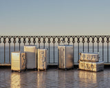 A collection of Monos aluminum suitcases in Aspen Silver in various sizes is arranged on a sunlit terrace with a wrought iron railing, overlooking the ocean in the background.