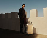 Adrien Brody wearing a dark coat and red scarf stands beside a white fortress-like wall, with a Monos aluminum carry-on plus in aspen silver in the sunlight. The deep blue sky contrasts with the shadows.