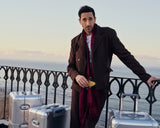 Adrien Brody leans on a wrought iron railing overlooking the ocean at dusk, surrounded by several Monos aluminum suitcases in aspen silver, holding a postcard in one hand.