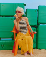 Woman in a striped swimsuit and yellow skirt eating ice cream on a beach with green suitcases in the background.